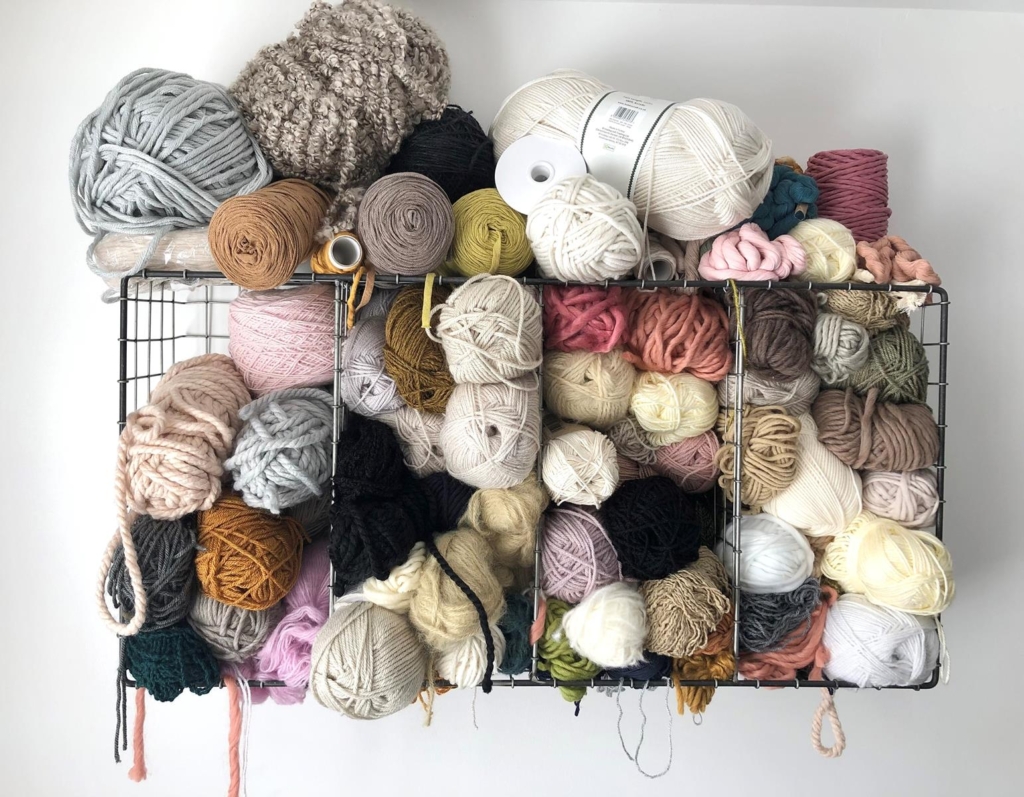 Wall rack holding dozens of balls of yarn in natural and pastel shades, in Weaveloveamy's studio workspace.