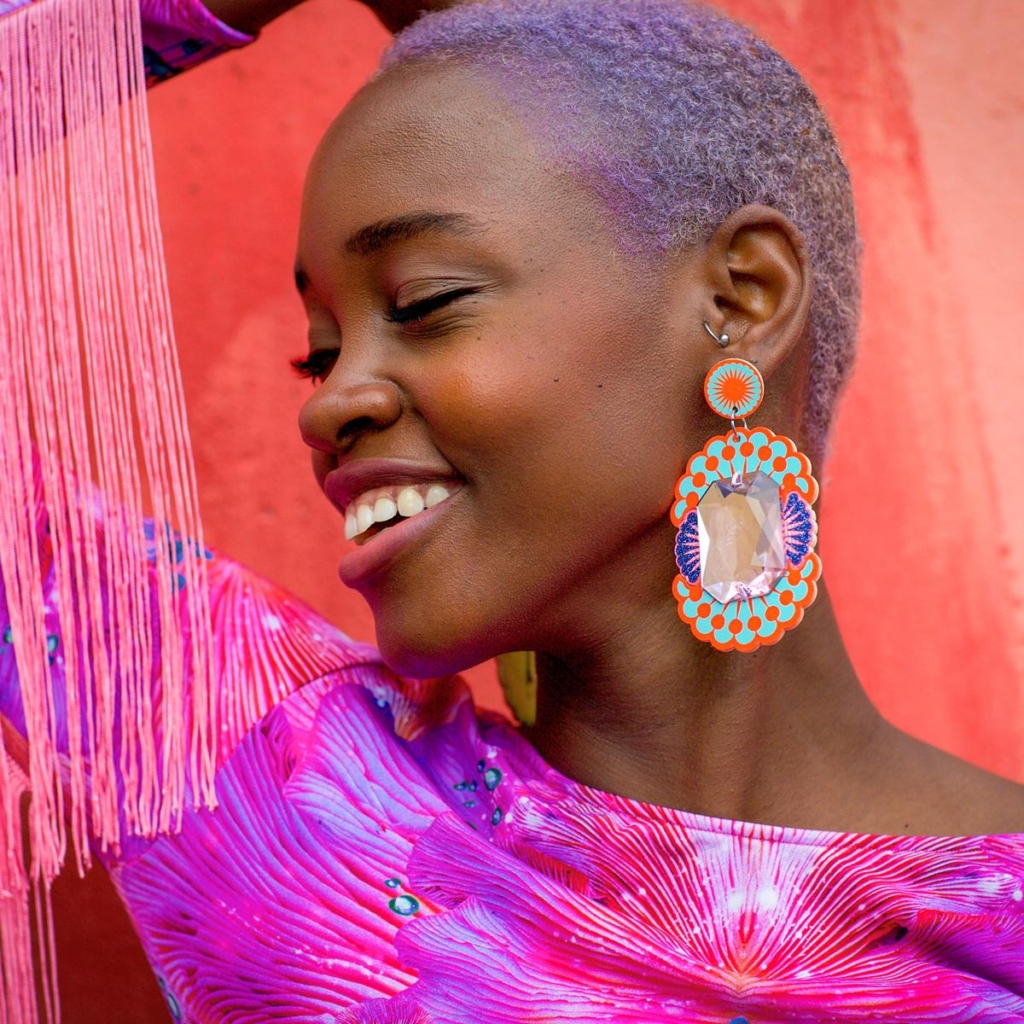 model wearing brightly coloured earrings in purples and pinks, with oversized clear 'gem' in the middle.