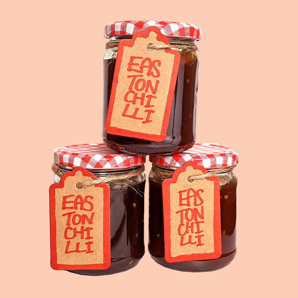 Three stacked jars of Easton Chilli Jam, with red and white gingham style tops, and 'Easton Chilli Jam' labels tied on with twine.