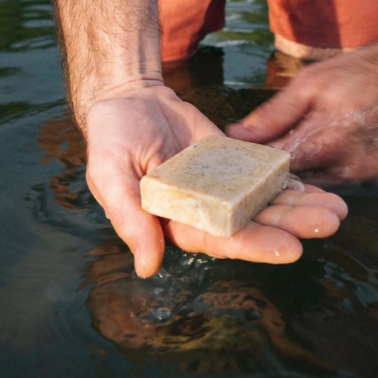 Natural soap with poppyseeds being used to wash hands in a shallow river, soap by Wylder Naturals.