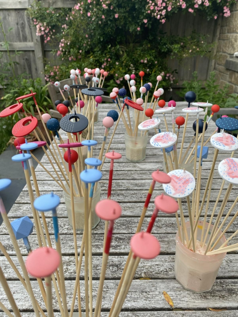 Around 50 large disc shaped wooden beads for earrings, in brighly painted beach and ice cream shades. They are drying outside on wooden skewers, on a wooden garden table. Made of sustainable wood, by Sea Pink Studio.