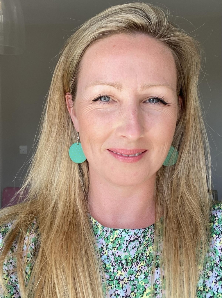 Headshot of Sally of Sea Pink Studio, dressed in shades of bright green and yellow. She is wearing matching green earrings - large, brightly painted wooden disc shapes.