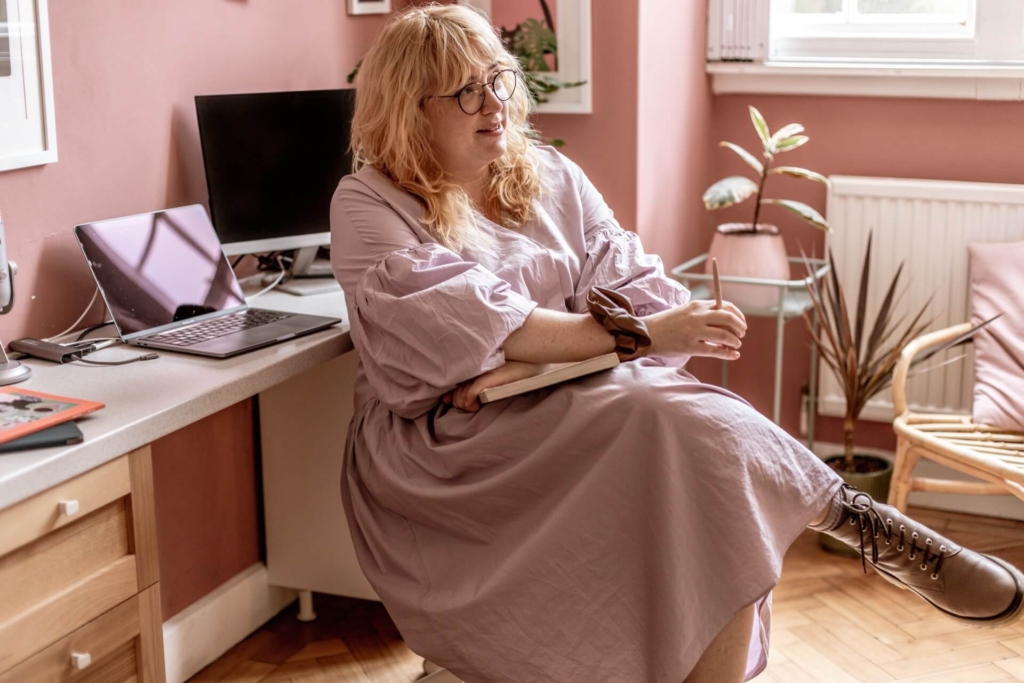 Aime Cox-Tennant sitting in a stylish office. She has curly light hair, rounded glasses, and is wearing a floaty tiered dress and beige lace up boots.