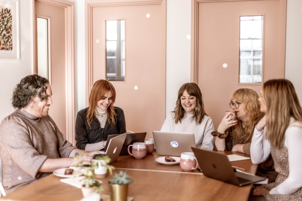 Four women and a man smiling while working at laptops around a large office desk. The room is decorated in stylish shades of pale salmon.