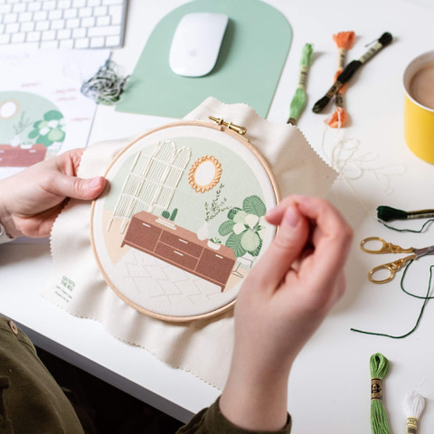 hands embroidering a scene with sideboard and plants, in a circular embroidery hoop