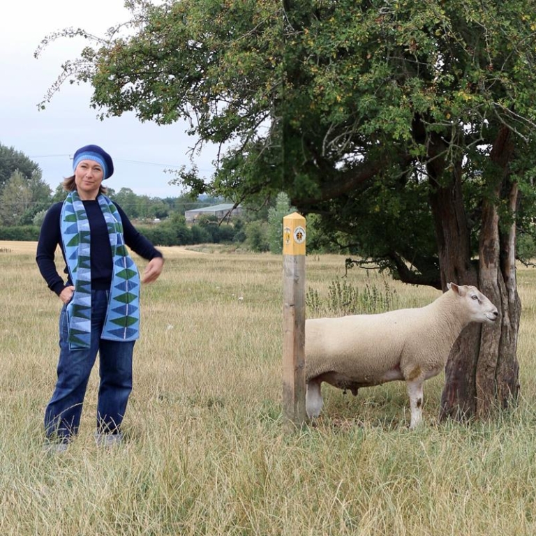 Anna Wheeler of Cotswold Knit standing in a field next to a sheep. She is wearing a light and dark blue long woollen scarf and hat set.