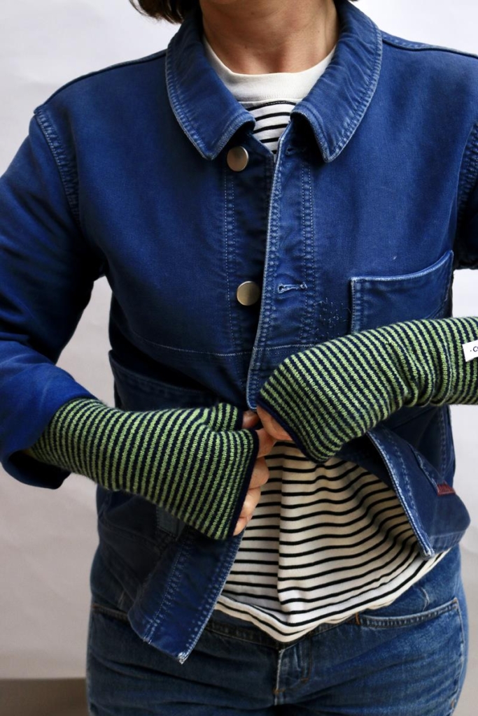 Model wearing 'Woodstock' knitted handwarmer fingerless gloves in 'Leaf' - shades of striped bottle green and navy.