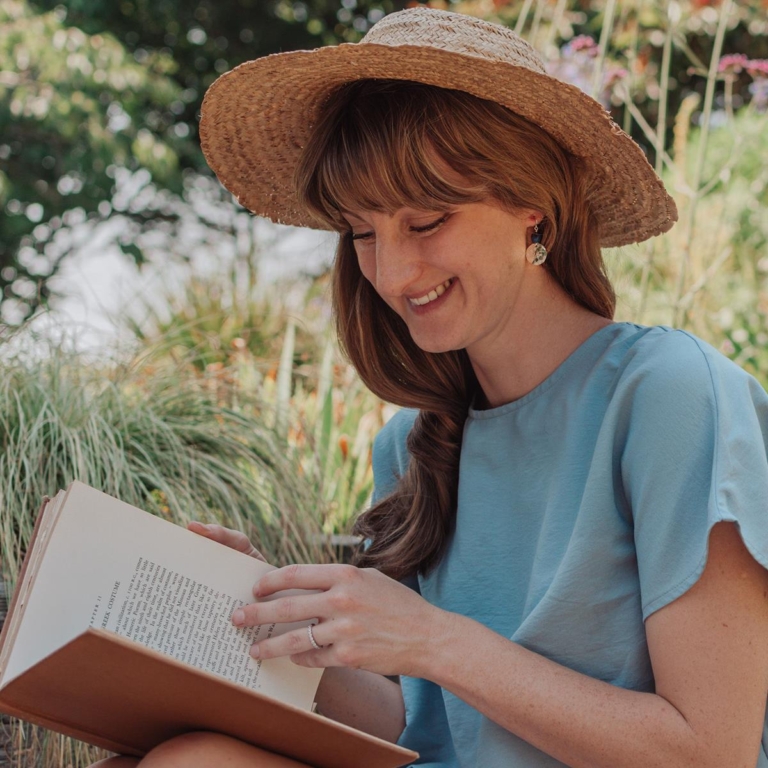 Nic Danning, founder of Nic Danning jewellery, sitting in a field wearing a straw hat, reading a book and smiling.