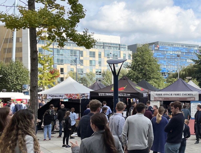 Scene at Temple Quay Market, Bristol, showing tented stalls outdoors on a paved area, with market goers in foreground.