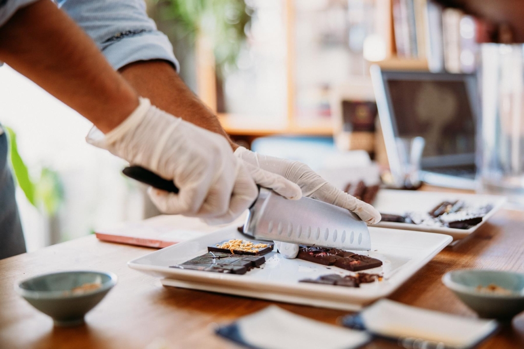 Chocolates being made by hand. Slabs of richly decorated chocolate are being cut with a large knive by gloved hands.