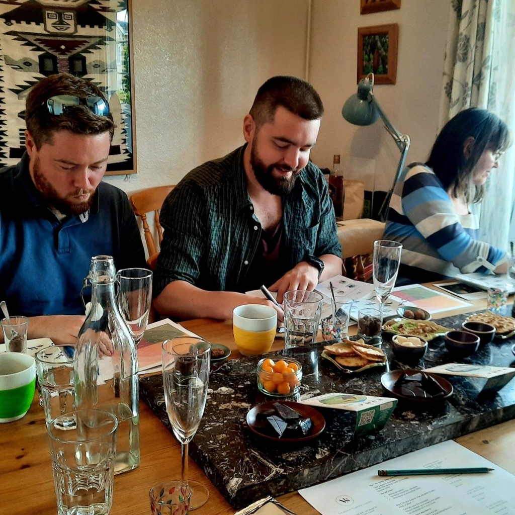 A group of people taking part in the Sensory Chocolate Tasting Experience at the home of Carlos Ayala. They are sitting around a table, smiling, with a selection of chocolates and ingredients in front of them. They have leaflets containing notes about the chocolate, and they are making notes.