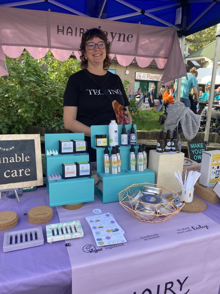 Hairy Jayne's market stall. Jayne is behind the stall, smiling, with her range of natural sustainable hair products, attractively packaged.