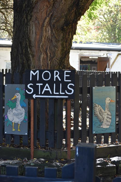 Large chalkboard sign saying 'More Stalls' on a wooden fence which is decorated with painted pictures of ducks.